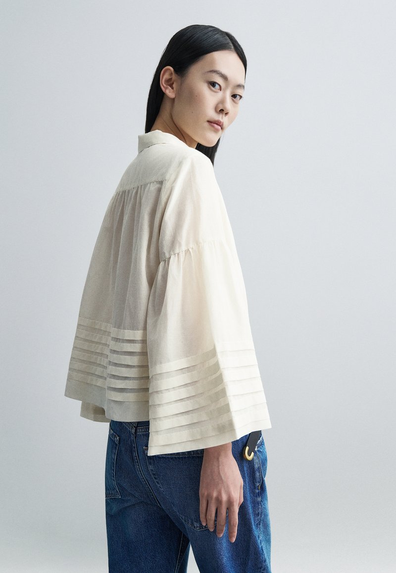 Ung kvinde med glat sort hår iført en løs, cremefarvet bluse med plisserede ærmer og blå jeans, der kigger over skulderen.