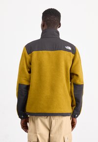 Giacca in pile con parte superiore grigio scuro e corpo giallo brillante. Presenta un colletto alto, chiusure a scatto sulle maniche e un logo sulla parte posteriore.