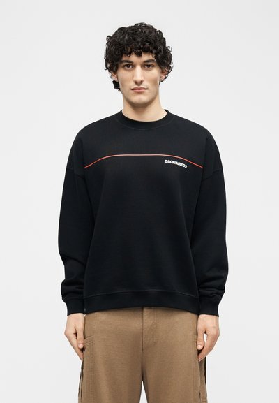 Jonge man met krullend haar die een zwart DSQUARED2-sweatshirt met rode bies en beige broek draagt, staand tegen een effen achtergrond.