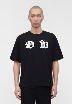 Mand med barberet hoved iført en sort oversized T-shirt med hvide abstrakte symboler på brystet, stående mod en ensfarvet lys baggrund.
