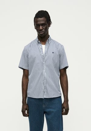 Tânăr în picioare purtând o cămașă albă cu mânecă scurtă, cu dungi înguste bleumarin, peste un tricou alb și blugi albaștri.