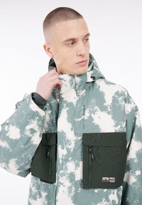 Hoodie aus einem grün-weißen Camouflage-Muster, ausgestattet mit zwei Reißverschlusstaschen, elastischen Bündchen und einem hohen Kragen.