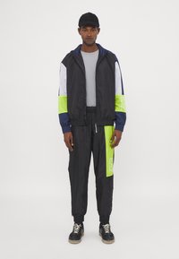 Black windbreaker with navy and lime green accents, paired with matching pants. Features a zip, elastic cuffs, and logo detail.