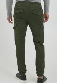 Man in olijfgroene cargobroek met elastische cuffs, flapzakken aan de zijkanten en achterkant, gecombineerd met zwarte sneakers en een grijs shirt met lange mouwen.