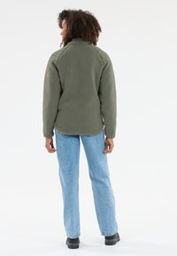 Vihreä fleece-takki, jossa on teksturoitu pinta ja korkea kaulus, yhdistettynä vaaleansinisiin suoriin farkkuihin ja mustiin jalkineisiin, nähtynä takaa.