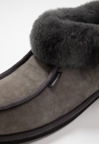 Zapatilla de ante gris con un suave puño de piel sintética de color gris oscuro. Presenta un borde cosido y una suela de goma negra. Etiqueta de la marca visible en el lateral.