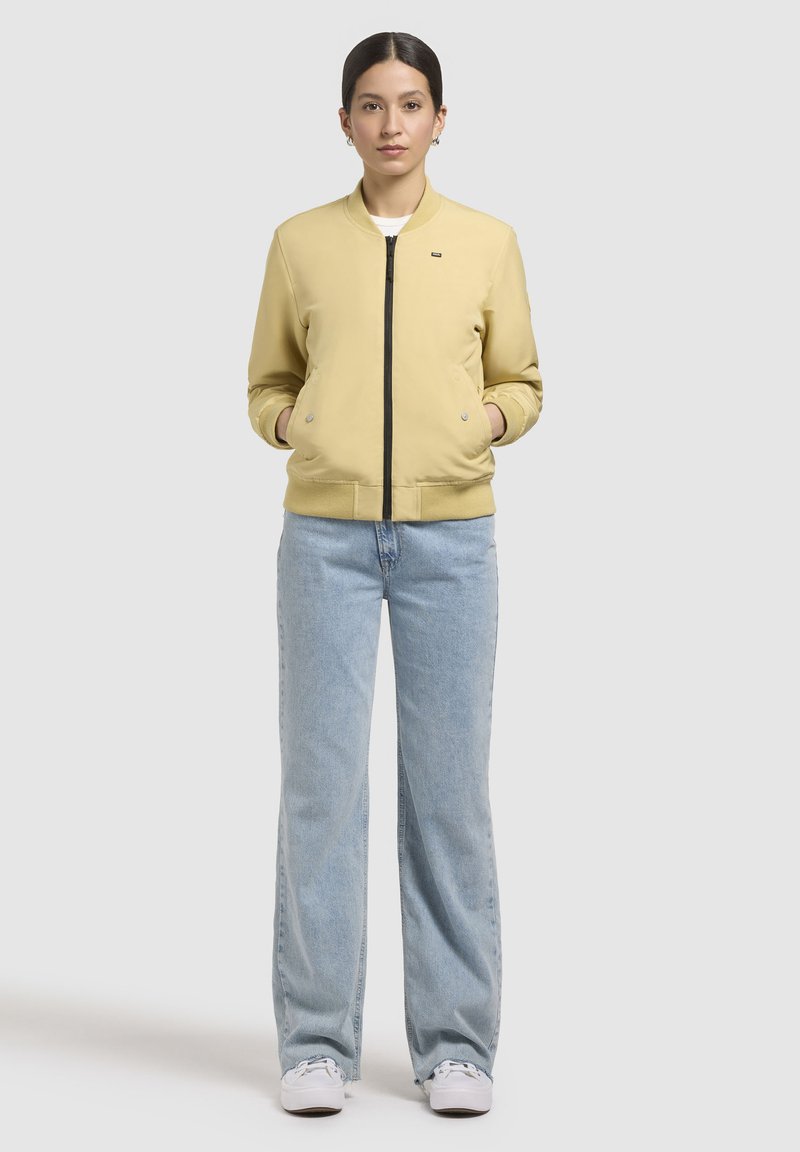 Gul bombarjacka med svart dragkedja, ribbade muddar och två sidofickor, matchad med ljusblå vida jeans och vita sneakers.