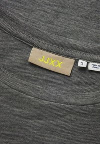 Tamno siva majica s okruglim izrezom, s bež etiketom na kojoj je žutim slovima napisano "JJXX" i veličina "S." Minimalni teksturirani detalji su vidljivi.