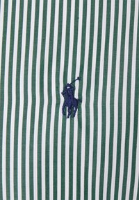 Zielono-biały materiał w paski o teksturowanym wykończeniu, z małym, haftowanym na ciemnoniebiesko logo konia z jeźdźcem.