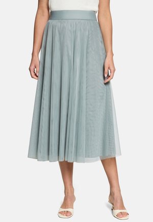 Kobieta ubrana w jasnoniebieską midi tiulową spódnicę z dopasowanym paskiem w pasie oraz białe sandały typu slide z odkrytymi palcami na białym tle.