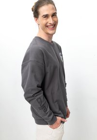 Grau Sweatshirt mit Rundhalsausschnitt, langen Ärmeln und Logo auf der Vorderseite und dem Ärmel. Glattes Material mit lockerer Passform.