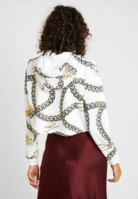 Žena s kudrnatými vlasy, otočená zády, oblečená v bílé zkrácené mikině s zlatým řetězem a logy Nike, doplněná burgundskou sukní.