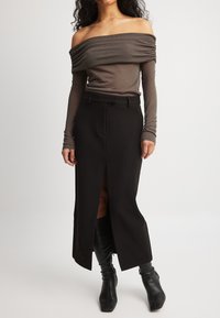 Kvinde iført en brun off-shoulder langærmet bluse, sort højtrævlet midi-nederdel med frontslids og sorte hælede støvler på en ensfarvet baggrund.
