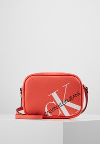 Röd syntetisk axelväska med rektangulär form, med en stor vit "CK"-logotyp och texten "CALVIN KLEIN JEANS". Inkluderar en avtagbar rem.