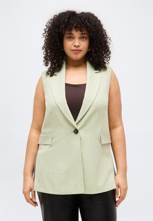 Kobieta o kręconych włosach ubrana w jasnozieloną, bezrękawnikową marynarkę na ciemnym topie i czarnych spodniach, stoi na tle jednolitego białego tła.