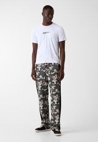 Weißes T-Shirt mit kurzen Ärmeln und "GUESS"-Logo, kombiniert mit grauer Camouflage-Hose mit abstrakten Mustern und schwarzen Seitenstreifen. Schwarze Sneaker.