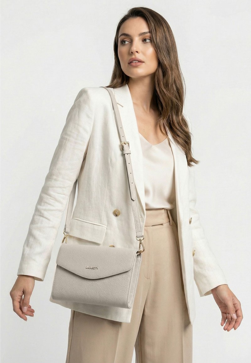 Mujer con pantalones beige y blazer blanco que lleva un bolso de hombro gris claro con correa ajustable y cierre de solapa.