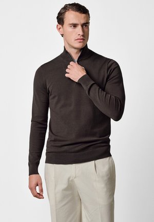 Uomo che sistema la cerniera di un maglione a collo alto marrone scuro, indossando pantaloni beige chiaro, guardando leggermente di lato su uno sfondo semplice.