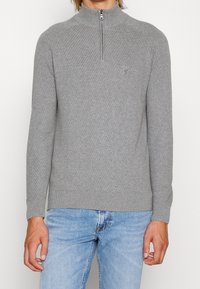 Grauer Reißverschluss-Pullover mit gerippter Textur und hohem Kragen. Mit einem kleinen Logo auf der Brust sowie geripptem Saum und Bündchen. Kombiniert mit blauen Jeans.