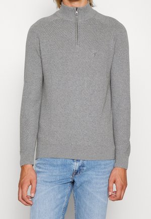 Grijze zip-up trui met geribbeld textuur en hoge kraag. Beschikt over een klein logo op de borst en geribbelde onderrand en manchetten. Gecombineerd met blauwe spijkerbroek.