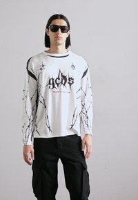 Camisa blanca de manga larga con estampado de alambre de espino negro, con el audaz logo negro "gcds" y el texto rojo "MILANO ITALY 1983". Pantalones cargo negros.