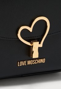 Μαύρη υφασμάτινη τσάντα χειρός με χρυσό κούμπωμα σε σχήμα καρδιάς και χρυσό λογότυπο "LOVE MOSCHINO". Κοντινή όψη που τονίζει το υλικό και τα μεταλλικά στοιχεία.
