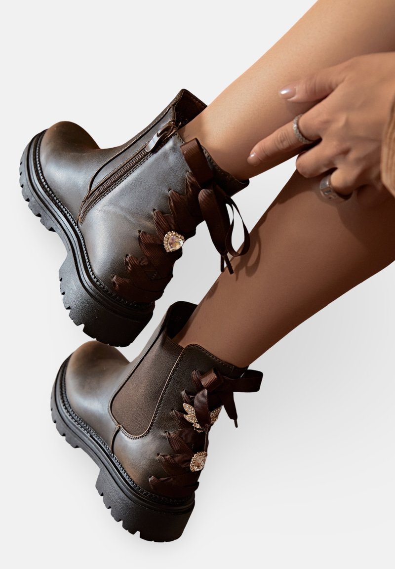 Bottines en cuir marron avec des rubans décoratifs et des accents en bijoux, portées sur des jambes avec des mains ajustant une bottine.