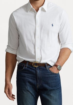 Mand iført hvid langærmet skjorte med lille blåt logo, mørkeblå jeans, brun læderbælte og brun ur med grøn urskive.
