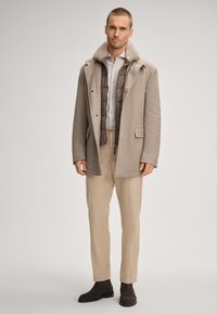 Jasny brązowy płaszcz z mieszanki wełny z szarym kołnierzem z baranka, noszony na ciemnobrązowej pikowanej kamizelce, w połączeniu z beżowymi spodniami i brązowymi butami.