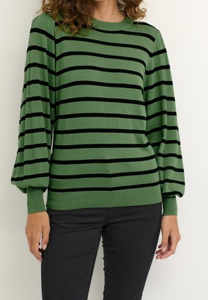 Kvinde iført en grøn sweater med vandrette sorte striber og sorte bukser, stående mod en ensfarvet lys baggrund.