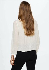 Blusa bianca testurizzata con maniche lunghe, caratterizzata da un taglio rilassato e orlo a goccia sul retro, abbinata a leggings neri.