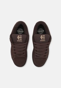 Pruunid suède tossud, padjakujulise keelega, lame paeltega ja kummist tallaga. "etnies" logo kuldse värviga keelega ja külge asetatud aktsentides.