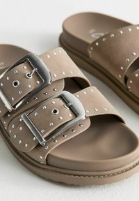Sandales marron avec deux larges brides ornées de clous en argent et de boucles métalliques réglables. Semelle lisse pour un confort optimal.