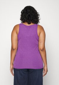 Lila ärmlös linne med rundad halsringning, tillverkat av ett texturerat tyg som ger lyster, bärs tillsammans med mörka denimbyxor.