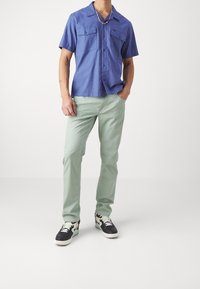 Camisa azul de manga corta con dos bolsillos en el pecho, combinada con pantalones verde claro y zapatillas negras con suela blanca y detalles verdes.