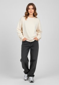 Felpa oversized beige con scollo rotondo e spalle scese, abbinata a jeans grigio scuro e sneakers bianche. Texture liscia visibile.