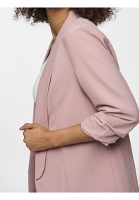 Světle růžový blazer vyrobený z hladké látky, s volnějším střihem, ohrnutými rukávy a jednou přední kapsou. Pohled z boku.