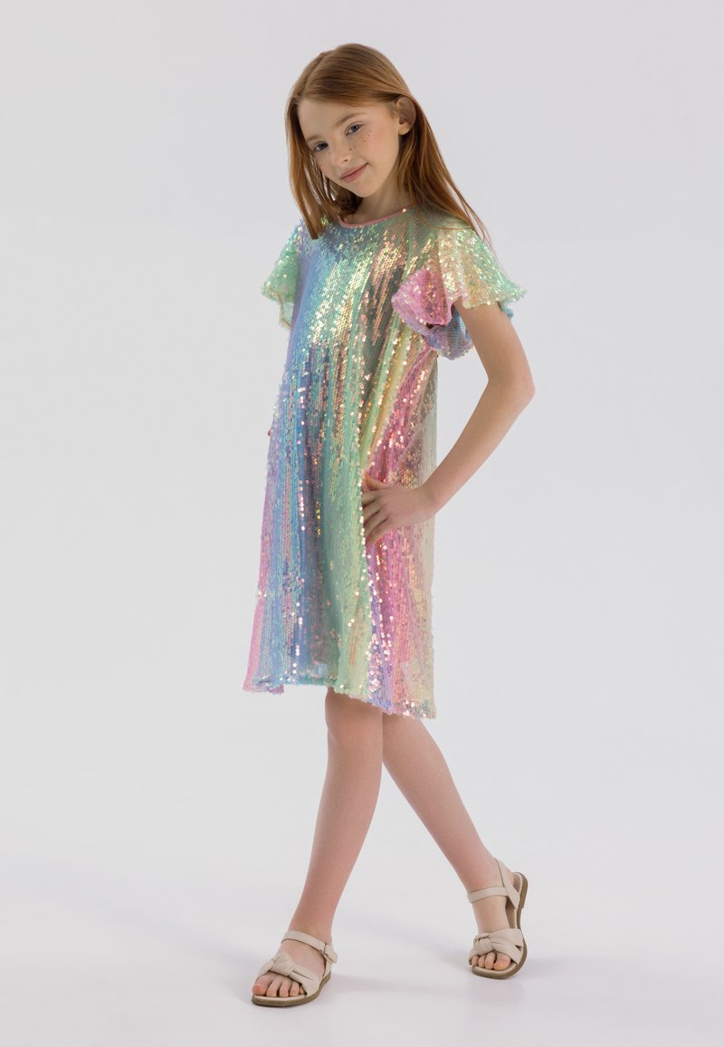 Robe en sequins irisés avec de courtes manches volantées, arborant un dégradé de couleurs arc-en-ciel. Le mannequin porte des sandales légères et pose avec une main sur la hanche.