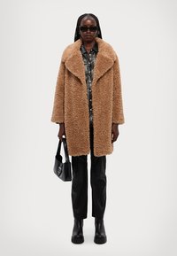 Cappotto oversized marrone e peloso con un ampio colletto, indossato sopra una camicia a fantasia. Pantaloni in pelle nera e stivaletti neri spessi completano il look.