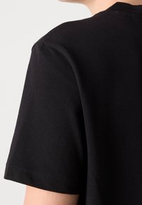 T-shirt noir en tissu texturé, col rond et manches courtes ; gros plan sur le dos révélant les détails des coutures et l'ajustement aux épaules.