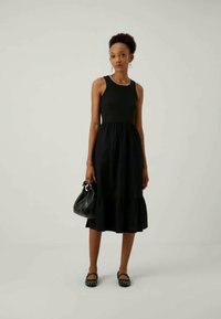 Robe noire sans manches avec corsage ajusté et jupe évasée, présentant un ourlet à volants. Portée avec des chaussures plates noires et un petit sac à main noir.