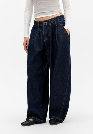 Personne portant un jean bleu foncé à jambes larges, un top blanc court à manches longues et des chaussures plates noires, une main dans la poche.