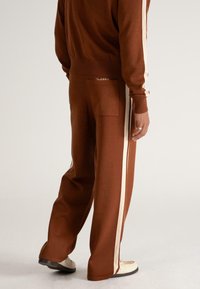 Bărbat îmbrăcat cu pantaloni sport maro cu dungi laterale albe și o jachetă asortată, stând pe un podea albă.