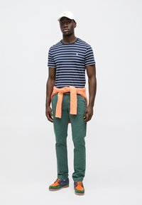 Navy striped t-shirt, green pants, orange sweater tied at the waist, and multicolored sneakers. Cotton and textured materials used.
