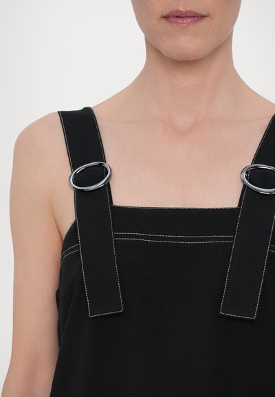 Crna majica s širokim naramenicama koja sadrži okruglu srebrnu metalnu opremu. Kontrastna bijela šavova duž rubova. Glatka, mat tekstura tkanine.