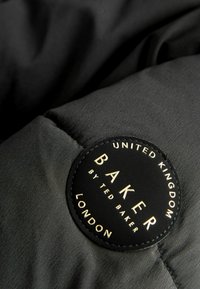 Tissu noir gonflé avec un patch en cuir noir rond portant une écriture dorée, étiqueté "BAKER par TED BAKER" et "ROYAUME-UNI LONDRES."