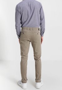 Chinos color marrón claro con un ajuste slim, bolsillos laterales y traseros, y cierre de botón. Combinados con una camisa de botón azul claro y zapatillas blancas.
