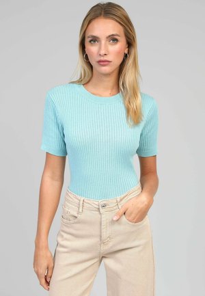 Femme aux cheveux blonds portant un haut côtelé bleu clair à manches courtes et un pantalon taille haute beige, une main dans la poche, fond neutre.