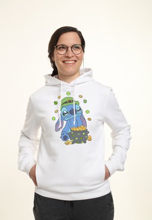 Uśmiechnięta kobieta w okularach i białej bluzie z kapturem, na której znajduje się postać z kreskówki w zielonym kapeluszu trzymająca garnek pełen złotych monet, otoczona koniczynami.