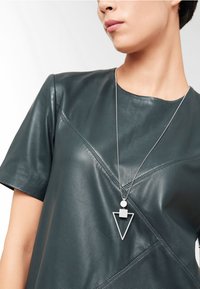 Teal lederen top met korte mouwen en een geometrische ketting met hanger, bestaande uit een driehoek en vierkant in zilver, tentoongesteld tegen een neutrale achtergrond.